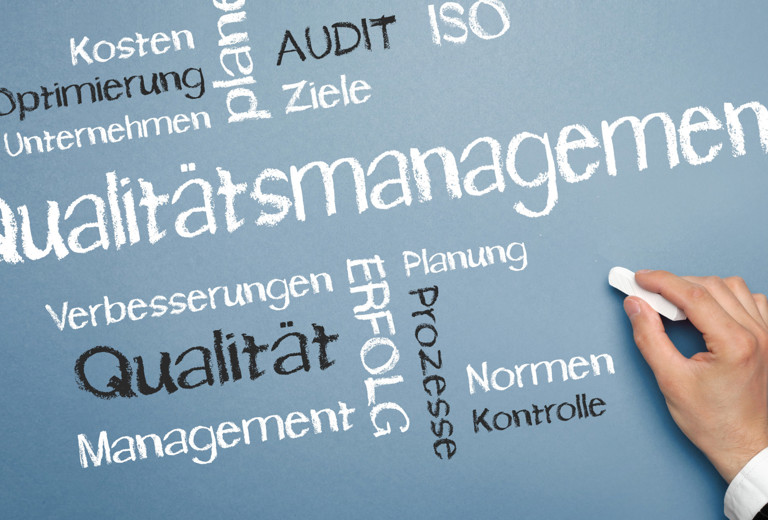Schreibtisch mit Taschenrechner, Stift und mit Quality Management beschriftetem Ordner